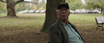 Movie still from “A Good Person” (2023), directed by Zach Braff – An older man wearing a hat sitting under a tree in a park; Close Up shot, Over the shoulder angle