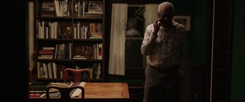 Movie still from “A Good Person” (2023), directed by Zach Braff – An older man standing in a living room with a book shelf in front of him; Medium shot, Low angle