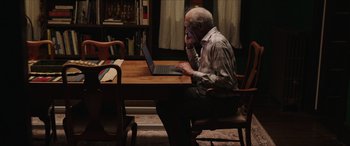 Movie still from “A Good Person” (2023), directed by Zach Braff – An older man sitting at a table using a laptop computer; Medium shot, High angle