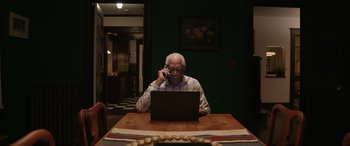 Movie still from “A Good Person” (2023), directed by Zach Braff – An older man sitting at a table using a laptop and talking on a cell phone; Medium shot, High angle