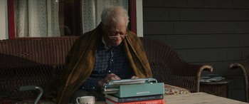Movie still from “A Good Person” (2023), directed by Zach Braff – An older man sitting at a table with a stack of books; Medium shot, Low angle