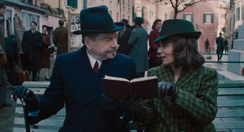 Movie still from “A Haunting in Venice” (2023), directed by Kenneth Branagh – A man and a woman wearing hats and holding an open book; Medium shot, Low angle