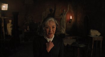 Movie still from “A Haunting in Venice” (2023), directed by Kenneth Branagh – An older woman in a black suit standing in front of a statue; Medium shot, Over the shoulder angle
