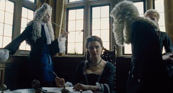 Movie still from “The Favourite” (2018), directed by Yorgos Lanthimos – A woman sitting at a table in front of two men in period garb; Medium shot, Over the shoulder angle