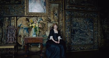 Movie still from “The Favourite” (2018), directed by Yorgos Lanthimos – A woman in a black dress sitting on a chair; Wide shot, High angle