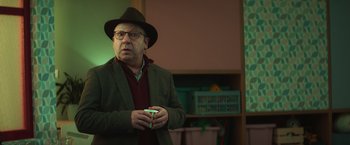 Movie still from “A Night at the Kindergarten” (2022), directed by Rafal Skalski – A man in a hat and glasses holding a cup in his hands; Medium shot, Over the shoulder angle
