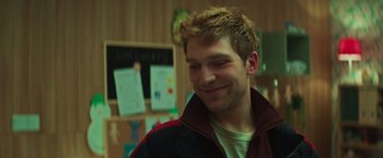 Movie still from “A Night at the Kindergarten” (2022), directed by Rafal Skalski – A young man smiling at the camera in front of a bulletin board; Close Up shot, Over the shoulder angle