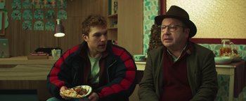 Movie still from “A Night at the Kindergarten” (2022), directed by Rafal Skalski – A man and a man sitting next to each other holding a plate; Medium shot, Over the shoulder angle