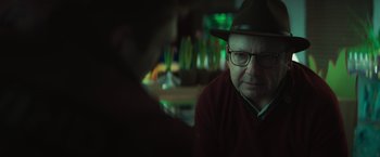 Movie still from “A Night at the Kindergarten” (2022), directed by Rafal Skalski – An older man wearing glasses and a hat is sitting at a table; Close Up shot, Over the shoulder angle