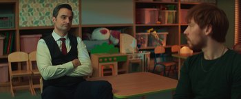 Movie still from “A Night at the Kindergarten” (2022), directed by Rafal Skalski – A man sitting at a wooden table in a room with toys; Medium shot, Over the shoulder angle