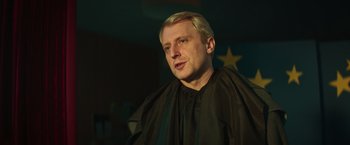Movie still from “A Night at the Kindergarten” (2022), directed by Rafal Skalski – A man with blonde hair wearing a black cape; Close Up shot, Over the shoulder angle