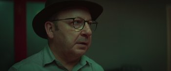 Movie still from “A Night at the Kindergarten” (2022), directed by Rafal Skalski – An older man wearing glasses and a hat; Close Up shot, Over the shoulder angle