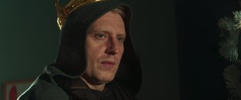 Movie still from “A Night at the Kindergarten” (2022), directed by Rafal Skalski – A man wearing a black robe and a gold hat; Close Up shot, Low angle