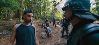 Movie still from “A Place to Fight For” (2023), directed by Romain Cogitore – A group of people standing in the woods near trees; Medium shot, Over the shoulder angle