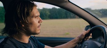 Movie still from “A Place to Fight For” (2023), directed by Romain Cogitore – A man sitting in the drivers seat of a car driving down a road; Close Up shot, Over the shoulder angle