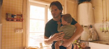 Movie still from “A Place to Fight For” (2023), directed by Romain Cogitore – A man holding a baby in his arms in a kitchen; Medium shot, Over the shoulder angle