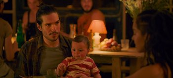Movie still from “A Place to Fight For” (2023), directed by Romain Cogitore – A man holding a baby in his arms in front of a table; Medium shot, Over the shoulder angle