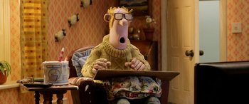 Movie still from “A Shaun the Sheep Movie: Farmageddon” (2019), directed by Will Becher – A man sitting in a chair with a wooden board in front of him; Medium shot, Over the shoulder angle