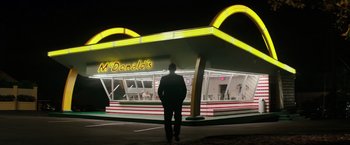 Movie still from “The Founder” (2016), directed by John Lee Hancock – A man standing in front of a mcdonald's restaurant at night; Extreme Wide shot, Low angle