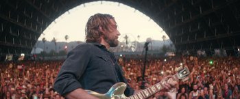Movie still from “A Star Is Born” (2018), directed by Bradley Cooper – A man with a beard playing an electric guitar in front of an audience; Medium shot, Over the shoulder angle