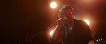Movie still from “A Star Is Born” (2018), directed by Bradley Cooper – A man holding a microphone in front of lights; Medium shot, Over the shoulder angle