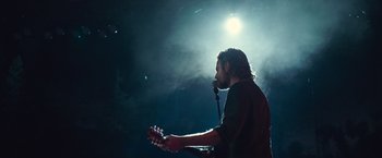 Movie still from “A Star Is Born” (2018), directed by Bradley Cooper – A man holding a microphone and playing a guitar; Medium shot, Low angle