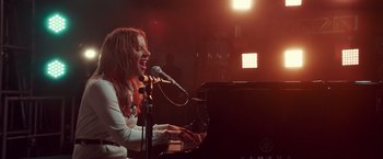 Movie still from “A Star Is Born” (2018), directed by Bradley Cooper – A woman singing into a microphone near a piano; Medium shot, Low angle