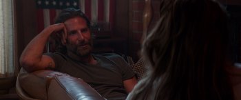 Movie still from “A Star Is Born” (2018), directed by Bradley Cooper – A man and a woman sitting on a couch in front of a flag; Medium shot, Over the shoulder angle
