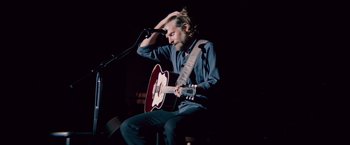 Movie still from “A Star Is Born” (2018), directed by Bradley Cooper – A man sitting on a chair playing a guitar; Medium shot, Low angle