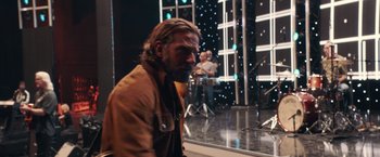Movie still from “A Star Is Born” (2018), directed by Bradley Cooper – A man in a brown jacket standing in front of a microphone; Medium shot, Over the shoulder angle