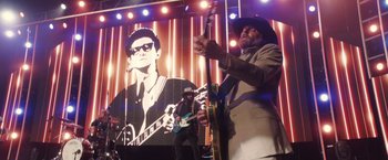 Movie still from “A Star Is Born” (2018), directed by Bradley Cooper – A man with a hat on playing guitar in front of a large picture of a man; Wide shot, Low angle