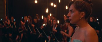 Movie still from “A Star Is Born” (2018), directed by Bradley Cooper – A group of people sitting in front of string lights holding instruments; Close Up shot, Over the shoulder angle