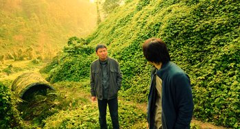 Movie still from “A Writer's Odyssey” (2021), directed by Yang Lu – Two men standing in front of a lush green hillside; Wide shot, High angle
