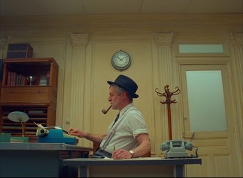 Movie still from “The French Dispatch” (2021), directed by Wes Anderson – A man with a pipe in his mouth writing on a table; Medium shot, Low angle