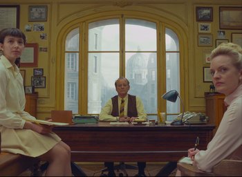 Movie still from “The French Dispatch” (2021), directed by Wes Anderson – A man sitting at a desk in front of a large window; Wide shot, Over the shoulder angle
