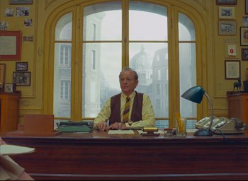 Movie still from “The French Dispatch” (2021), directed by Wes Anderson – An older man sitting at a desk in front of a window; Wide shot, Low angle