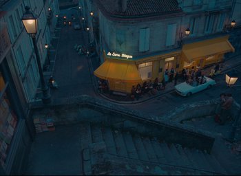 Movie still from “The French Dispatch” (2021), directed by Wes Anderson – An aerial view of a street corner at night; Extreme Wide shot, High angle