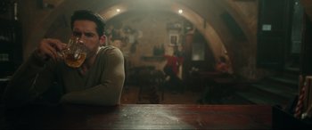 Movie still from “Accident Man: Hitman's Holiday” (2022), directed by George Kirby – A man sitting at a table in front of a bar; Medium shot, Over the shoulder angle