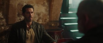 Movie still from “Accident Man: Hitman's Holiday” (2022), directed by George Kirby – A man in a leather jacket sitting in front of a brick wall; Medium shot, Over the shoulder angle