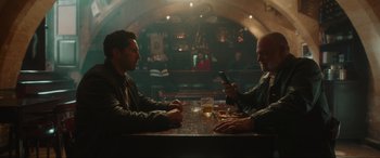 Movie still from “Accident Man: Hitman's Holiday” (2022), directed by George Kirby – Two men sitting at a table in front of a cell phone; Wide shot, Over the shoulder angle