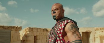 Movie still from “Accident Man: Hitman's Holiday” (2022), directed by George Kirby – A man with a face tattoo is standing in front of a stone wall; Close Up shot, Low angle