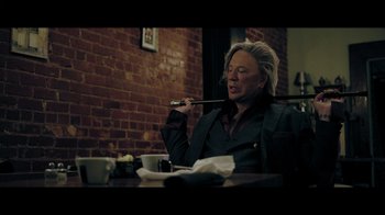 Movie still from “Adverse” (2020), directed by Brian A. Metcalf – A man sitting at a table with a pipe in his mouth; Medium shot, Low angle