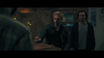 Movie still from “Adverse” (2020), directed by Brian A. Metcalf – A man sitting at a table in front of a bottle of alcohol; Medium shot, Over the shoulder angle