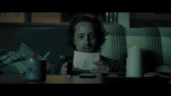 Movie still from “Adverse” (2020), directed by Brian A. Metcalf – A man sitting at a table holding a piece of paper in front of his face; Close Up shot, Low angle