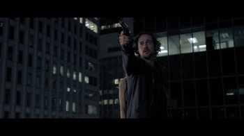 Movie still from “Adverse” (2020), directed by Brian A. Metcalf – A man holding a gun while standing in front of a building; Medium shot, Low angle