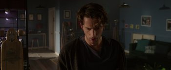 Movie still from “Afterlife of the Party” (2021), directed by Stephen Herek – A man with long brown hair is standing in a dark room; Close Up shot, Over the shoulder angle