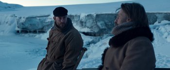 Movie still from “Against the Ice” (2022), directed by Peter Flinth – A man and a woman sitting on a chair in the snow; Medium shot, Over the shoulder angle