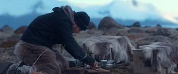 Movie still from “Against the Ice” (2022), directed by Peter Flinth – A man in a black jacket and a black hat is cooking food on an outdoor stove; Wide shot, High angle