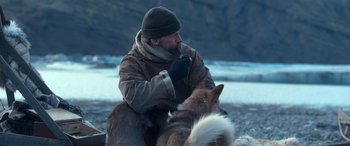 Movie still from “Against the Ice” (2022), directed by Peter Flinth – A man sitting next to a dog on the beach; Medium shot, High angle