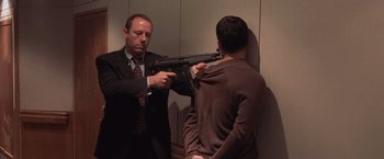Movie still from “Air Force One” (1997), directed by Wolfgang Petersen – A man in a suit and tie holding a machine gun up to the back of a man; Medium shot, Over the shoulder angle